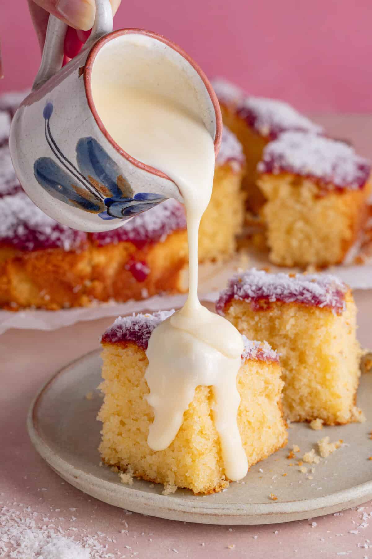 A piece of jam and coconut cake on a plate with custard being poured over it.