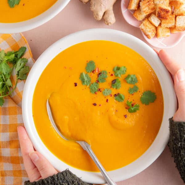 Two hands holding a bowl of carrot ginger and chilli soup.